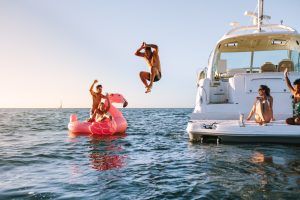 A white vessel anchored at sea. Two people sit on the boat deck, while another jumps next to a person floating on a giant flamingo.
