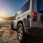 The back of a white Jeep Wrangler on a muddy rocky trail beside a shallow stream on a sunny day with blue skies.
