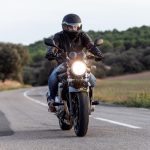 A man wearing a helmet, leather jacket, and safety gloves while riding a motorcycle on an open road.