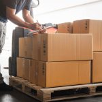A warehouse worker writes on a clipboard as he inspects a shipment of supplies. The supplies are on top of a wooden palette.