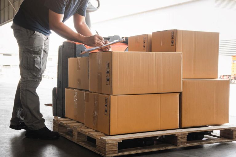 A warehouse worker writes on a clipboard as he inspects a shipment of supplies. The supplies are on top of a wooden palette.