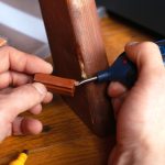 A closeup on a pair of hands using a small handheld tool to repair the leg of a chipped piece of furniture.