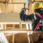 A construction worker wearing a hard hat, safety gloves, harness, and tool belt while installing roof trusses.