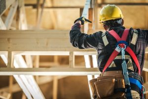 A construction worker wearing a hard hat, safety gloves, harness, and tool belt while installing roof trusses.