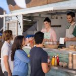 Three customers stand at a food truck counter decorated with bunting. Two vendors serve drinks and snacks from inside.