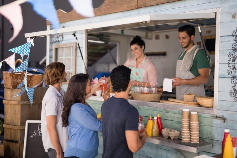 Three customers stand at a food truck counter decorated with bunting. Two vendors serve drinks and snacks from inside.