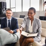 Four business people sitting in a well-appointed office. Two of them are shaking hands after making a deal.