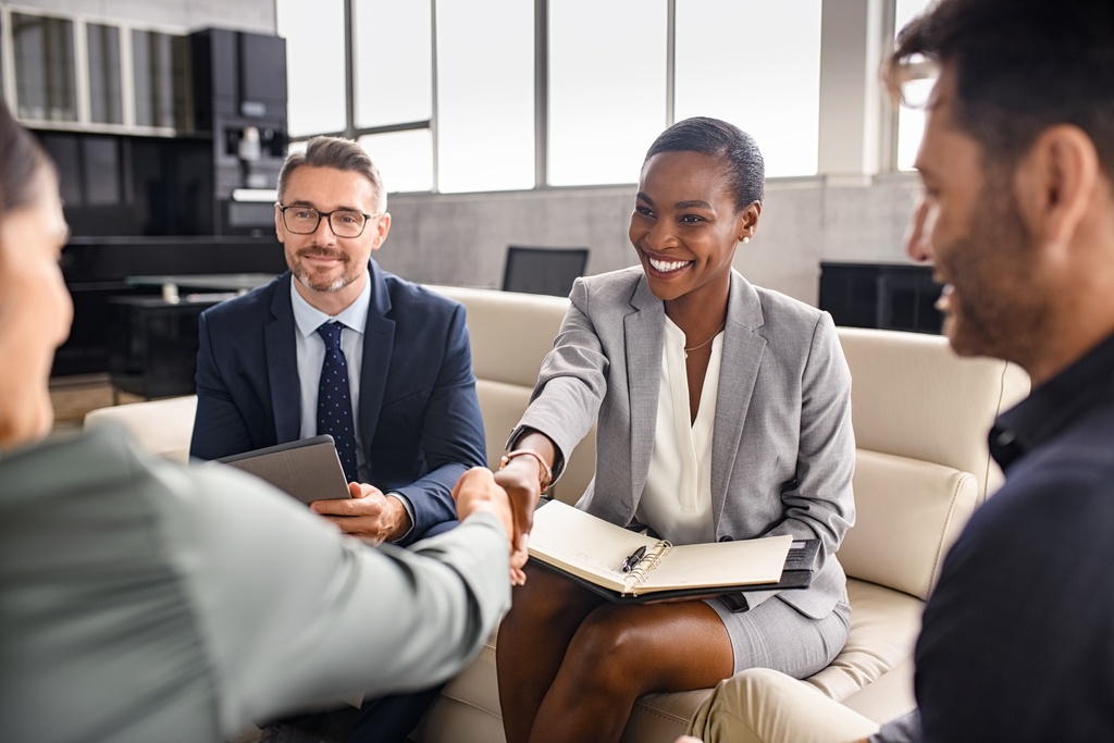 Four business people sitting in a well-appointed office. Two of them are shaking hands after making a deal.