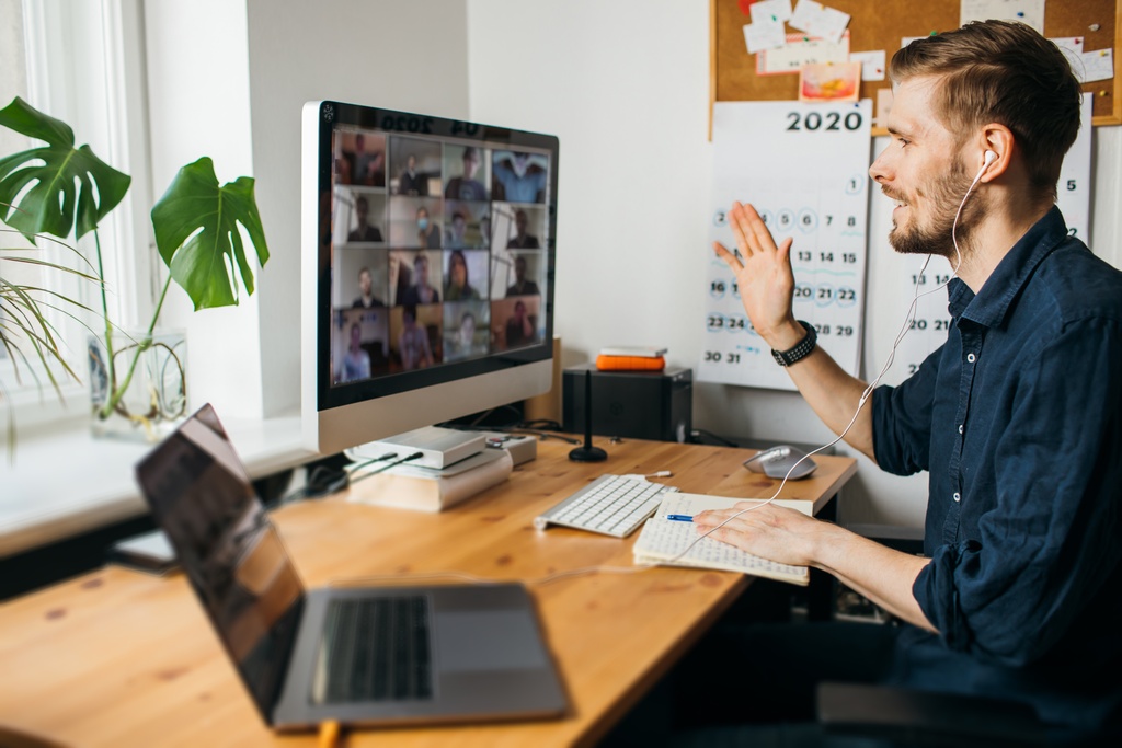 A man sitting at a desk with a laptop and computer screen. He is waving at the people on the video call on the screen.