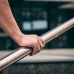 A close-up of a person holding a rounded silver handrail. Stairs and a building are blurred in the background.