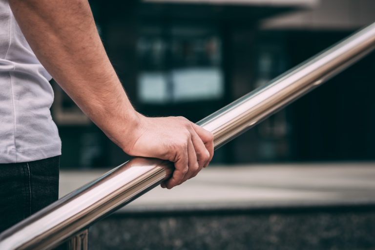 A close-up of a person holding a rounded silver handrail. Stairs and a building are blurred in the background.