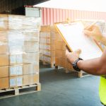 A worker in a high-visibility vest writes on a clipboard. He stands near palletized cardboard boxes wrapped in plastic.