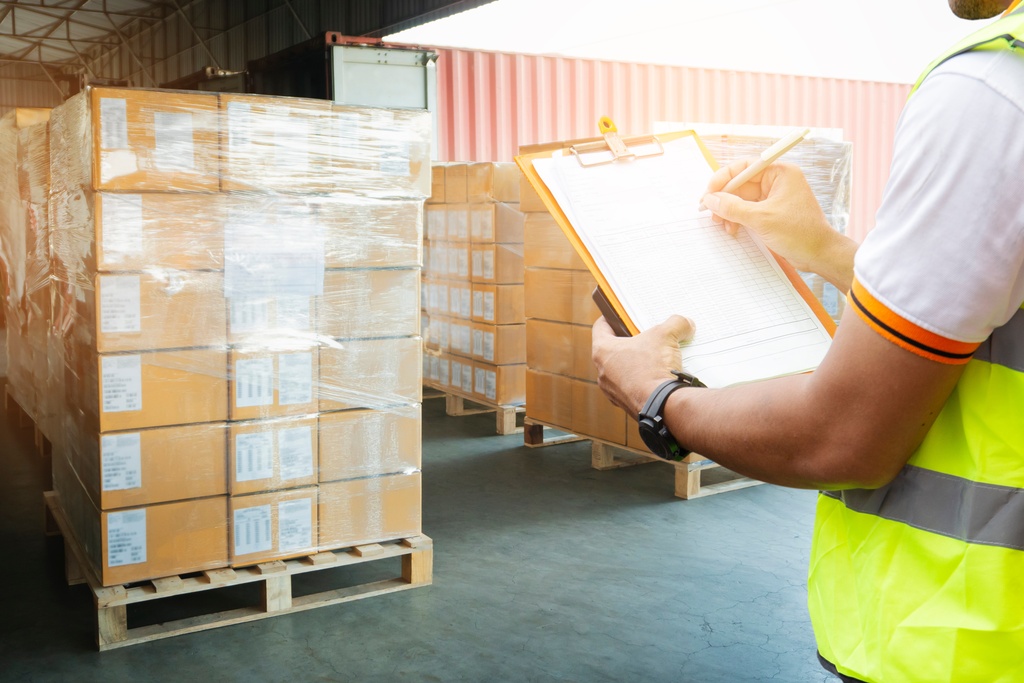 A worker in a high-visibility vest writes on a clipboard. He stands near palletized cardboard boxes wrapped in plastic.
