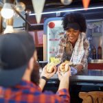 Smiling food vendor leans from a truck window, handing two hot dogs to a customer at an outdoor counter.