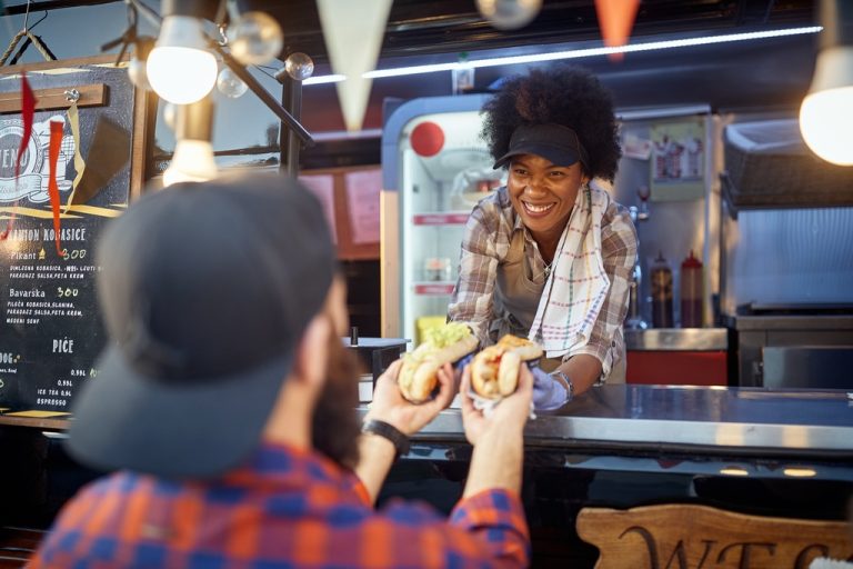 Smiling food vendor leans from a truck window, handing two hot dogs to a customer at an outdoor counter.