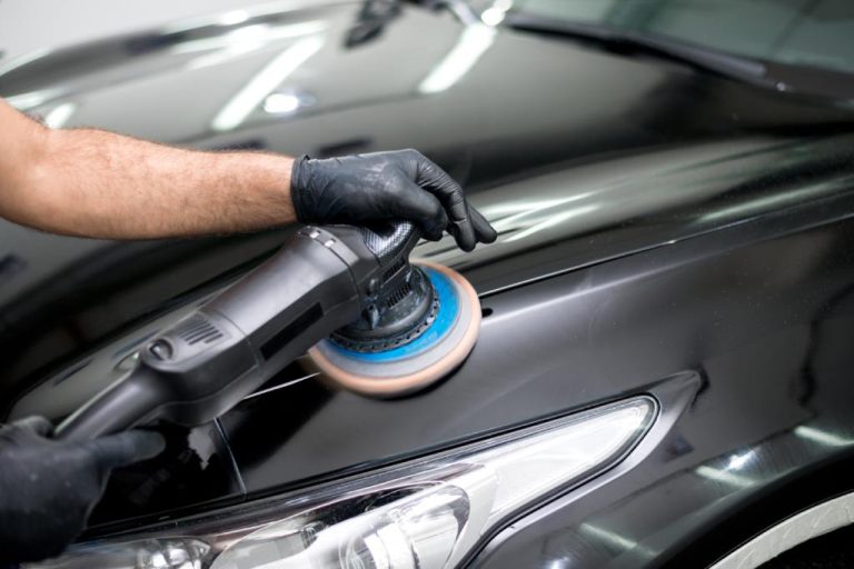 A man wearing a black glove is using a machine on a car’s surface. The car is black and highly reflective.