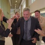 A group of people are standing in a hallway. They are all gathered around an older man with gray hair.