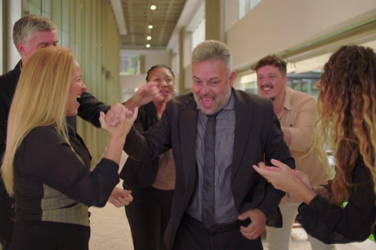A group of people are standing in a hallway. They are all gathered around an older man with gray hair.