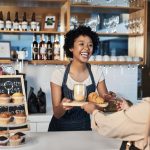 A woman wearing an apron stands behind the counter of a café. She hands a customer a tray with a coffee and a croissant.