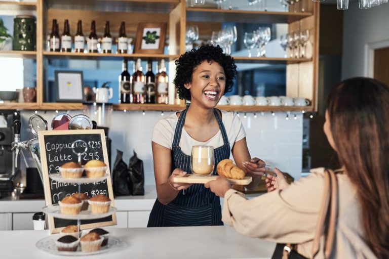 A woman wearing an apron stands behind the counter of a café. She hands a customer a tray with a coffee and a croissant.