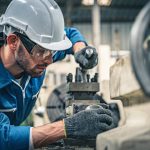 A man with beard stubble is wearing a hard hat and is leaning into a mechanical device.