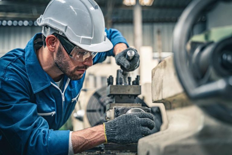 A man with beard stubble is wearing a hard hat and is leaning into a mechanical device.