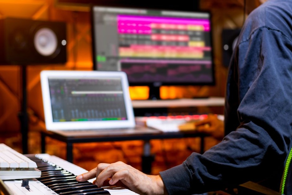 A musician sits at his desk with a computer monitor, a laptop, and a mixing table on the lefthand side of him.