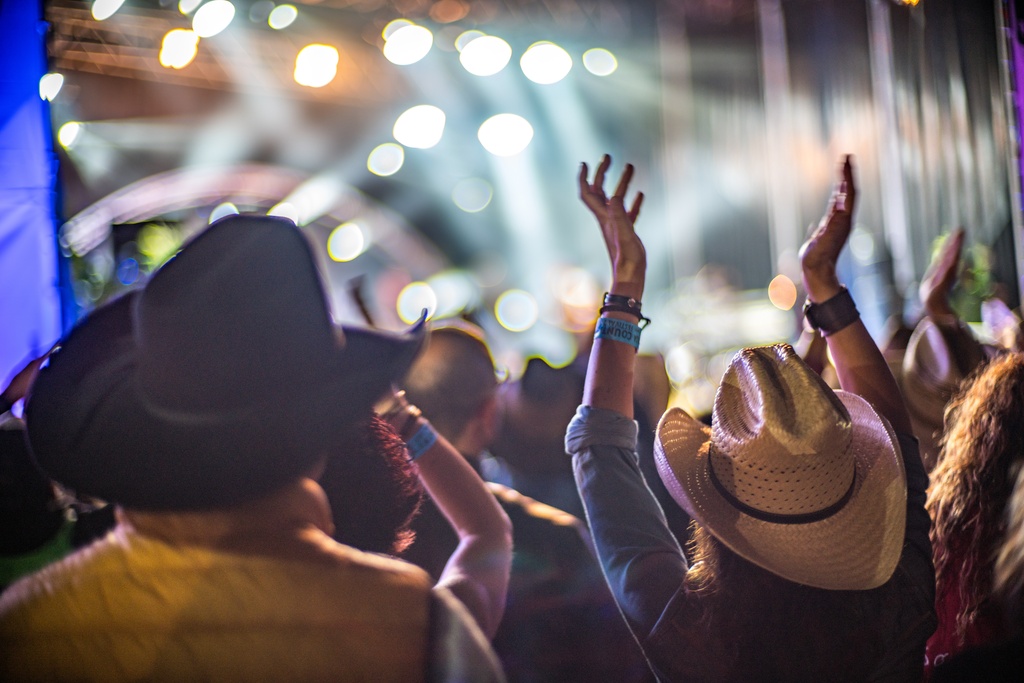 A crowd of people wearing cowboy hats raises their hands while enjoying live music at a brightly lit concert.