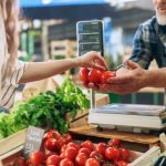 A man at a produce stand handing a woman a few bright red tomatoes. He has a scale sitting in front of him.