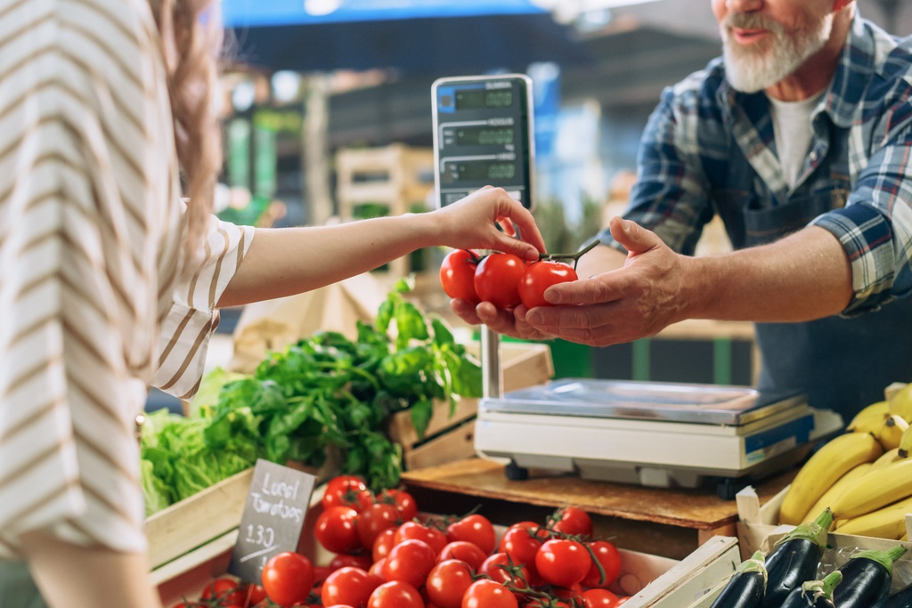 A man at a produce stand handing a woman a few bright red tomatoes. He has a scale sitting in front of him.