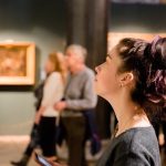 A close-up of a woman with her brown hair in a messy bun looking up at an exhibit in an art gallery or museum.