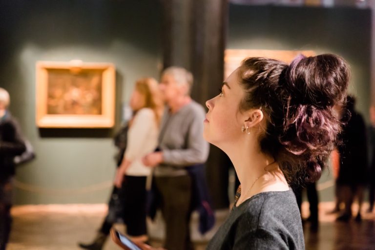 A close-up of a woman with her brown hair in a messy bun looking up at an exhibit in an art gallery or museum.