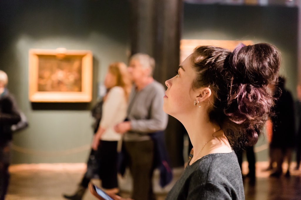 A close-up of a woman with her brown hair in a messy bun looking up at an exhibit in an art gallery or museum.