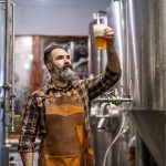 A bearded man in an apron holding a glass of amber beer and examining it inside a craft brewery with tanks behind him.