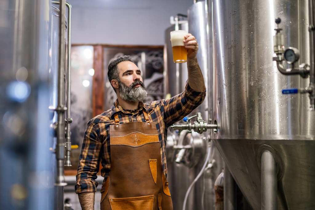 A bearded man in an apron holding a glass of amber beer and examining it inside a craft brewery with tanks behind him.
