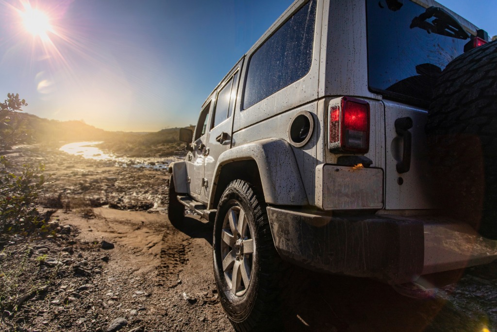 A Jeep Wrangler parked on a rocky dirt trail. A small stream flows through the rocks ahead with the sun reflecting off the water.