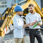 A builder and sales consultant standing on an outdoor lot reviewing documents while looking at heavy construction machinery.