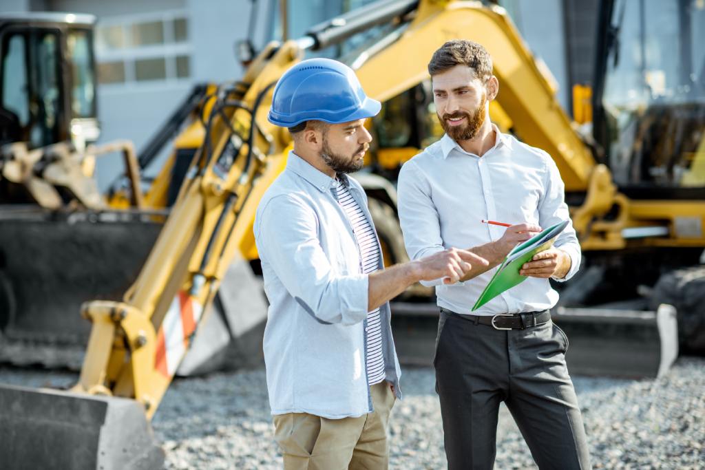 A builder and sales consultant standing on an outdoor lot reviewing documents while looking at heavy construction machinery.