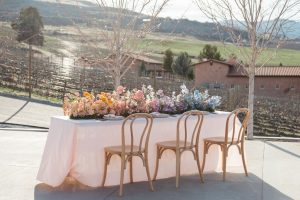 A wedding table outside next to a vineyard. The table is covered in a pink tablecloth and many colorful flowers.