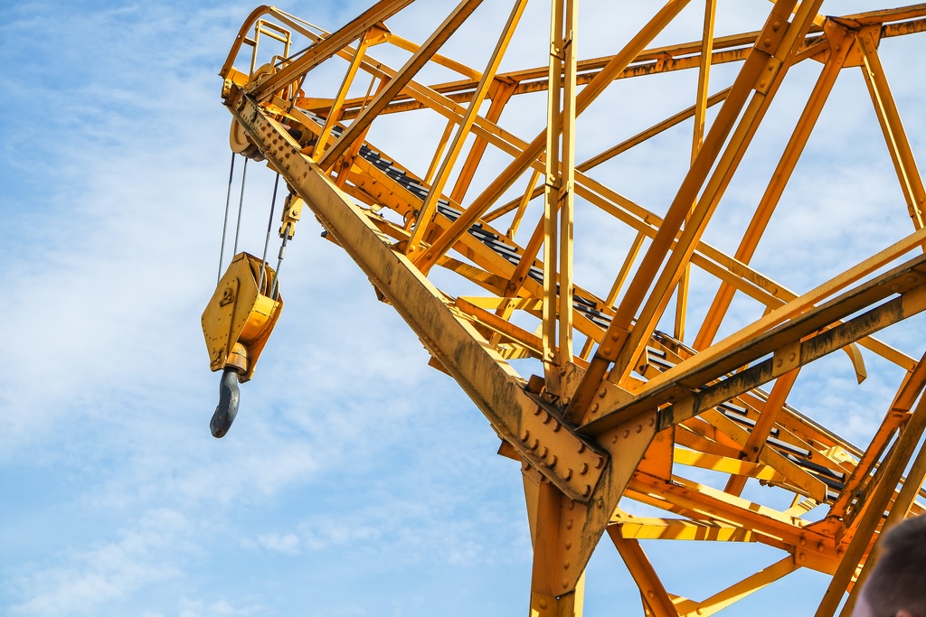 A tall yellow construction crane with a long boom and lifting hook extending upward against a clear blue sky.