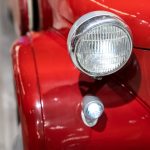 A close-up of the rear light and chrome trim on a shiny red vintage car, showing curved bodywork against a blurred background.