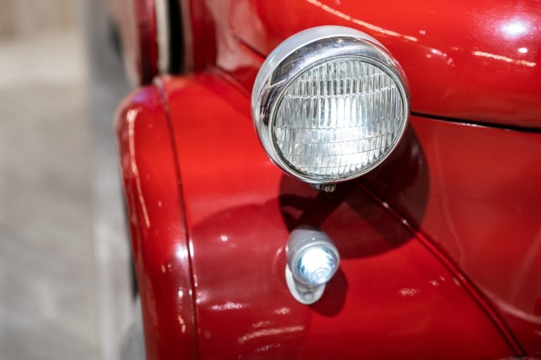 A close-up of the rear light and chrome trim on a shiny red vintage car, showing curved bodywork against a blurred background.