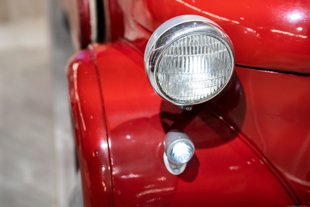 A close-up of the rear light and chrome trim on a shiny red vintage car, showing curved bodywork against a blurred background.