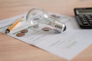 A printed electricity bill on a desk beside a glowing light bulb, scattered coins, a calculator, and a pen.