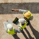Three contractors stand together on a jobsite in high-vis safety vests and hard hats. One holds a computer.