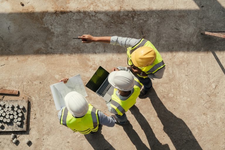 Three contractors stand together on a jobsite in high-vis safety vests and hard hats. One holds a computer.
