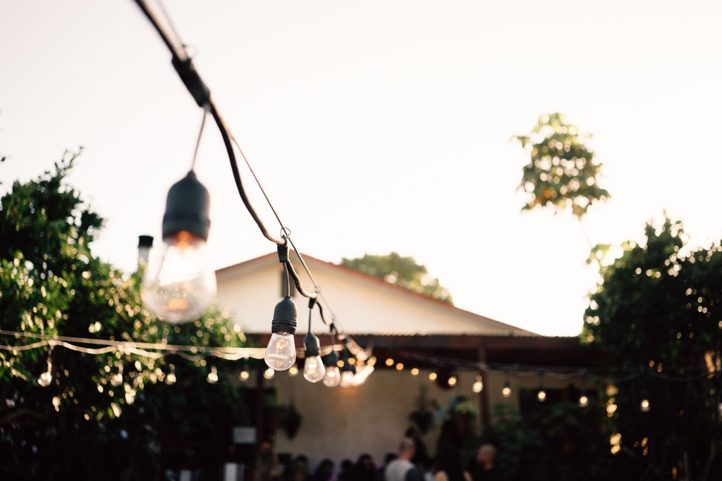 String lights hang overhead in a backyard at sunset, glowing warmly above a small gathering near a house.
