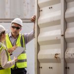 Two industrial workers wearing hard hats and safety vests talking outside a white shipping container on a work site.