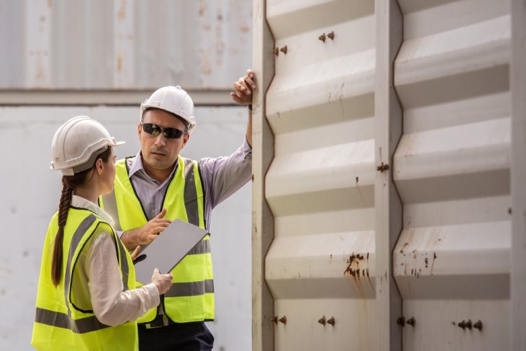 Two industrial workers wearing hard hats and safety vests talking outside a white shipping container on a work site.