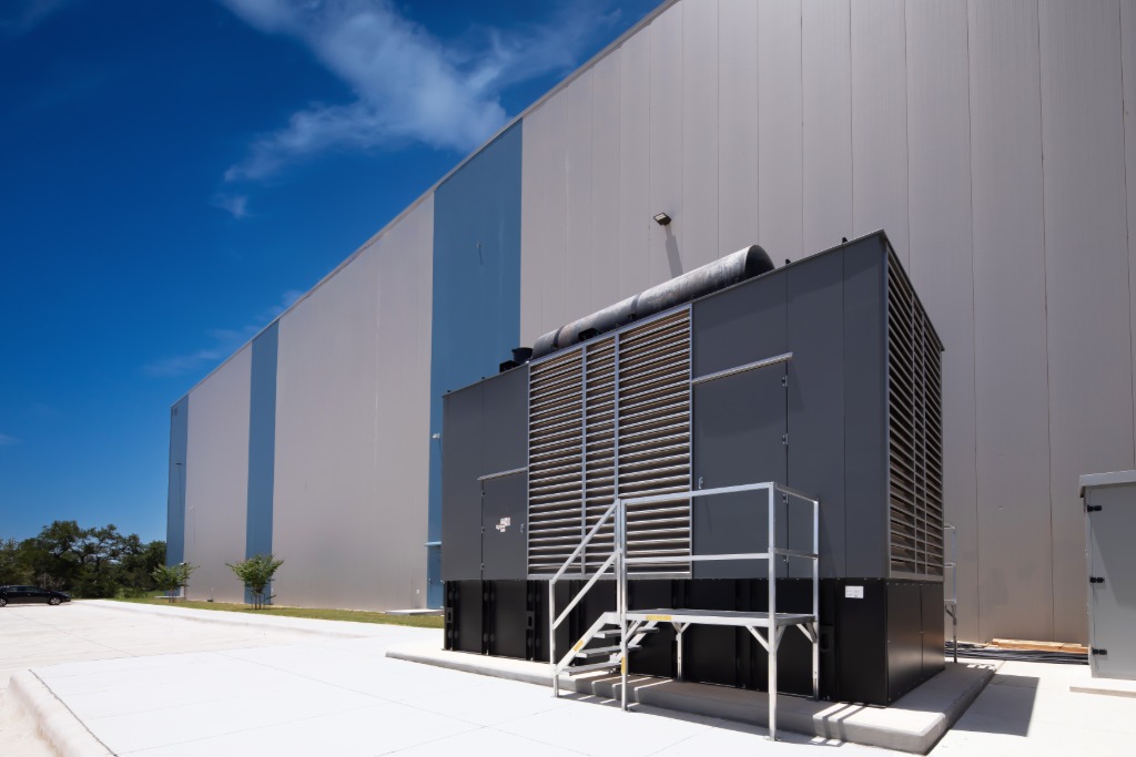 A large black backup power generator installed outside during the daytime next to a large industrial facility.